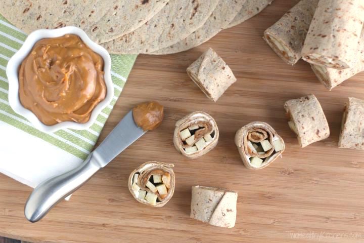 Assembled apple and peanut butter pinwheels on a wood cutting board next to a knife and a bowl of peanut butter.