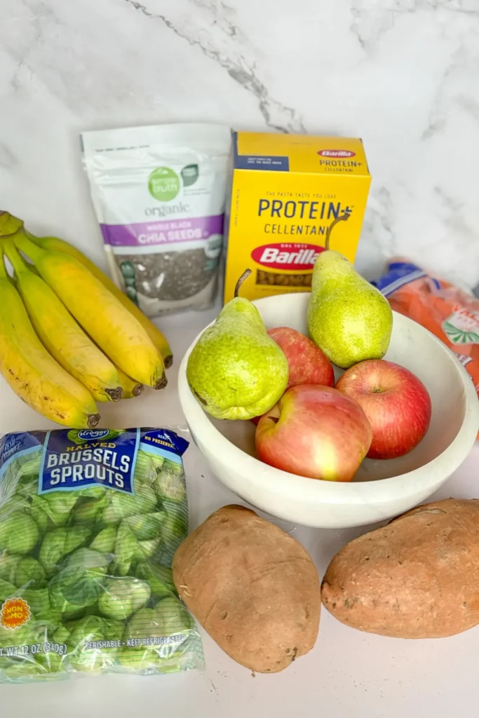 A fruit bowl with apples and pears in it on a counter next to sweet potatoes, Brussel sprouts, bananas, pasta and chia seeds.