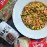 A bowl of rice, edamame, and veggies next to a bag of long grain rice, a can of black beans and a bag of lentils.