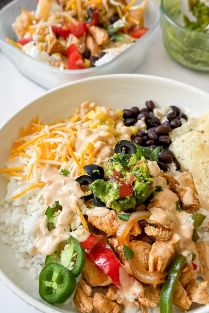 A fully assembled chicken fajita bowl served over rice with shredded cheese, guacamole and jalapenos in a bowl on a counter next to a bowl of guacamole.