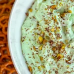 A close-up of the side of a large bowl of pistachio protein fluff topped with chopped pistachios on a tray with pretzel twists.