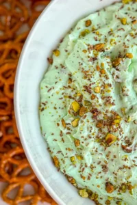 A close-up of the side of a large bowl of pistachio protein fluff topped with chopped pistachios on a tray with pretzel twists.