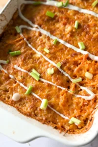 A close-up of the buffalo chicken quinoa casserole in a white casserole dish drizzled with ranch dressing and topped with green onions.