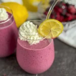 A lemon berry smoothie garnished with lemon wedges and whipped cream on a counter with lemons in the background.