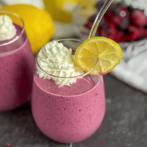 A lemon berry smoothie garnished with lemon wedges and whipped cream on a counter with lemons in the background.