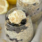 Two small glasses with blueberry lemon cheesecake overnight oats garnished with whipped cream and blueberries on a counter next to lemons.