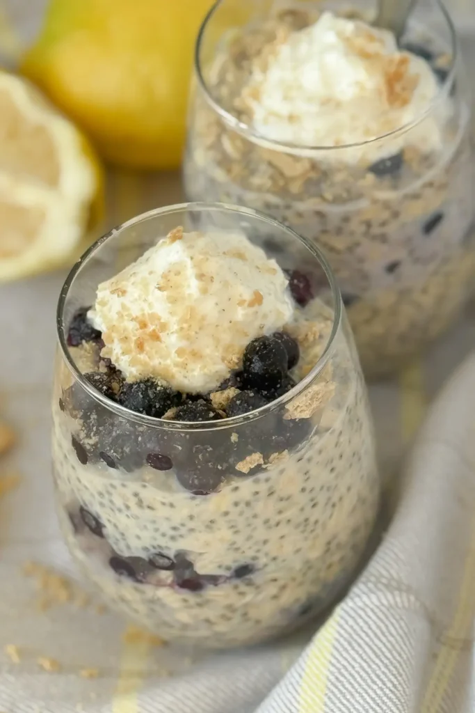Two small glasses with blueberry lemon cheesecake overnight oats garnished with whipped cream and blueberries on a counter next to lemons.