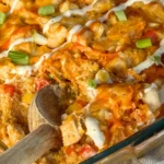 A baked buffalo chicken quinoa casserole drizzled with green onions and ranch dressing with a wooden spoon digging into it.