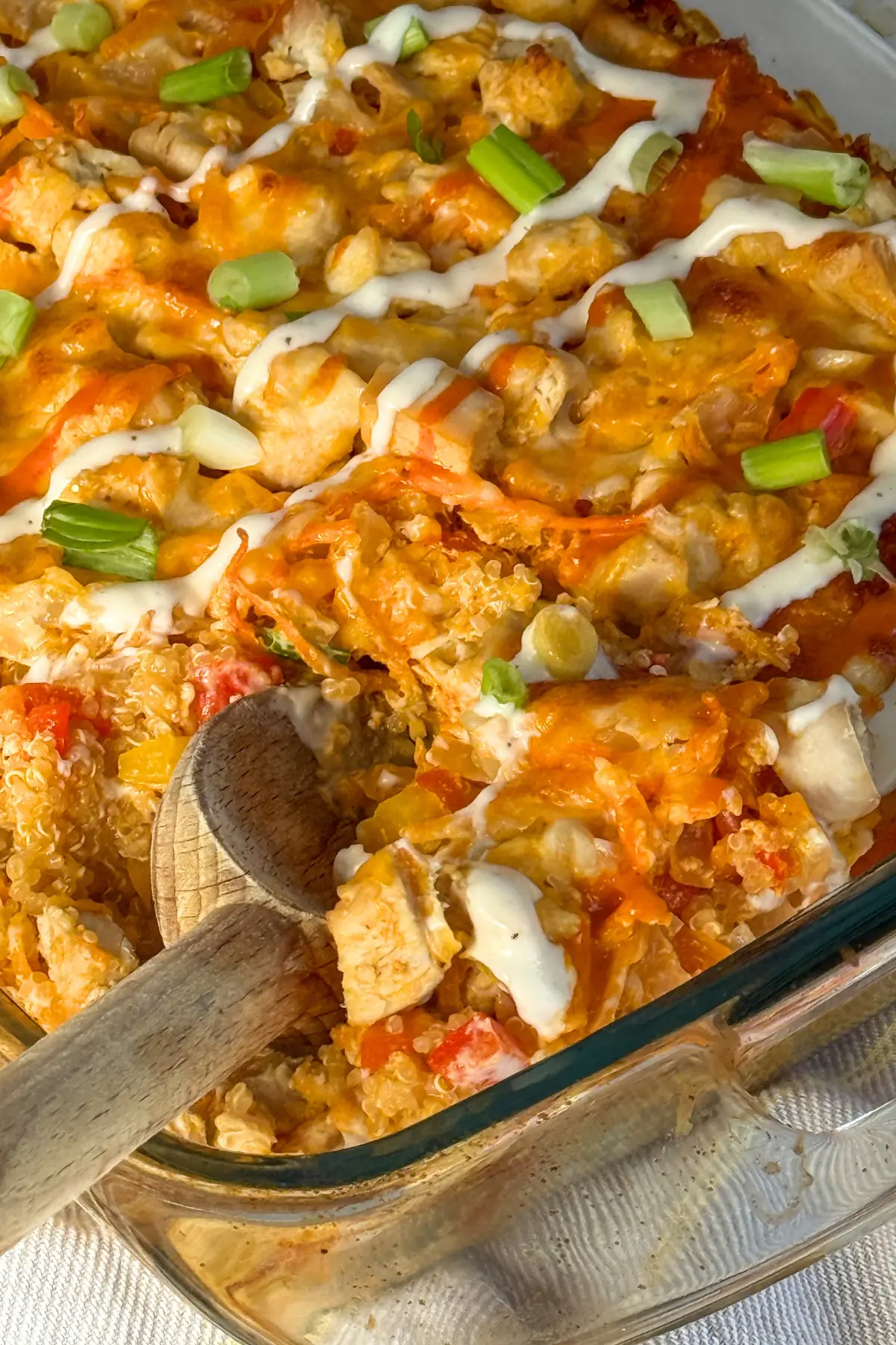 A baked buffalo chicken quinoa casserole drizzled with green onions and ranch dressing with a wooden spoon digging into it.