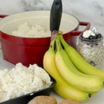 A pot of rice next to a storage container of cooked and cooled rice, a bunch of green bananas, two potatoes and overnight oats in a jar on a counter.