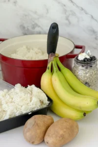 A pot of rice next to a storage container of cooked and cooled rice, a bunch of green bananas, two potatoes and overnight oats in a jar on a counter.