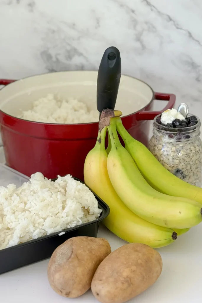 A pot of rice next to a storage container of cooked and cooled rice, a bunch of green bananas, two potatoes and overnight oats in a jar on a counter.