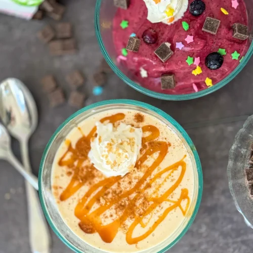 A birds-eye view of two bowls of soft serve; one with frozen banana and caramel and the other frozen berries with dark chocolate chips and whipped cream.