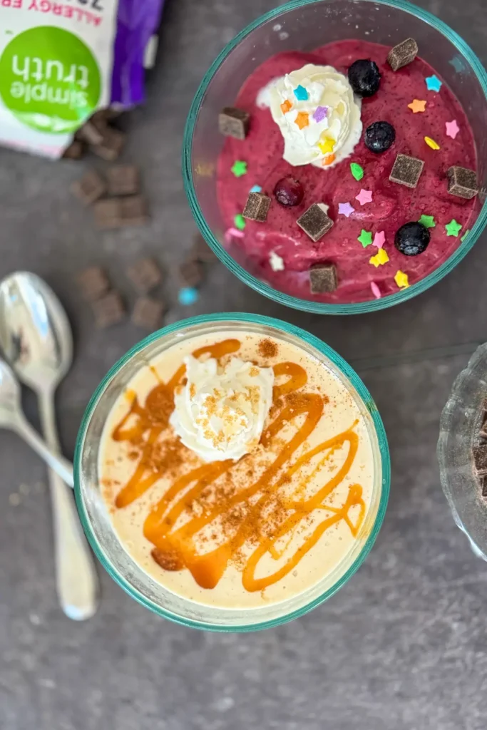 A birds-eye view of two bowls of soft serve; one with frozen banana and caramel and the other frozen berries with dark chocolate chips and whipped cream.