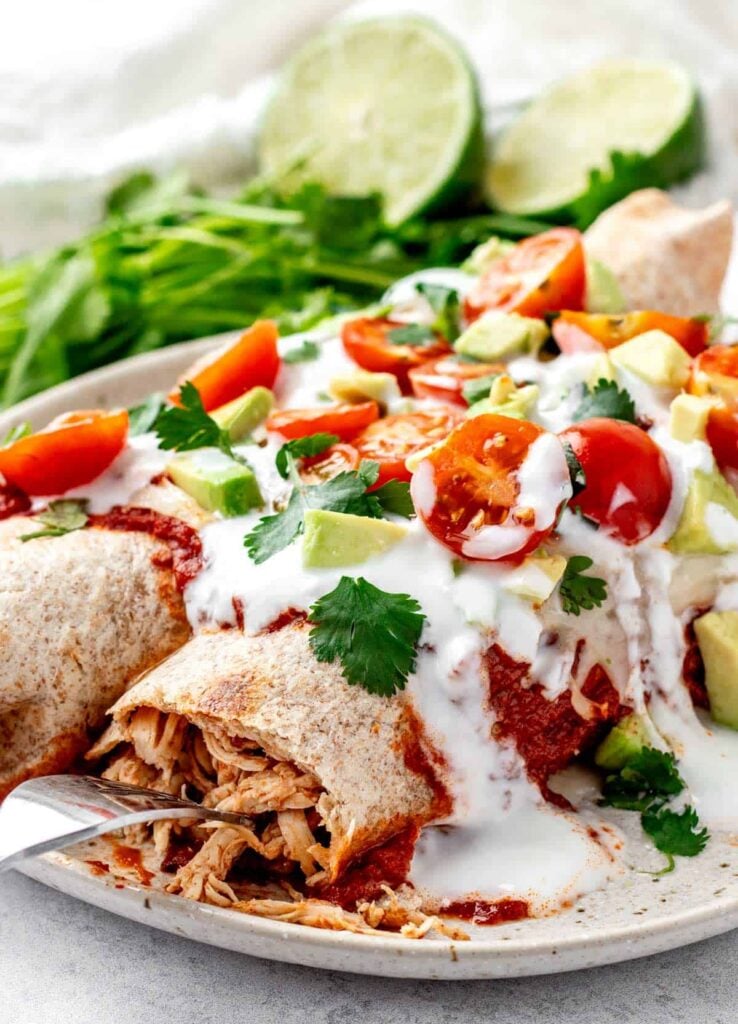 Two healthy chicken enchiladas on a plate topped with sour cream and tomato slices, garnished with cilantro.