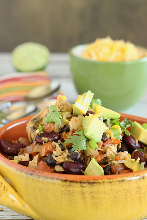A bowl of three bean chicken chili topped with avocado slices on a table.