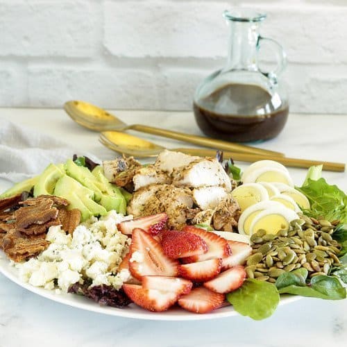 A low carb, high fiber cobb salad with strawberries assembled on a plate sitting on a counter next to a carafe of dressing.