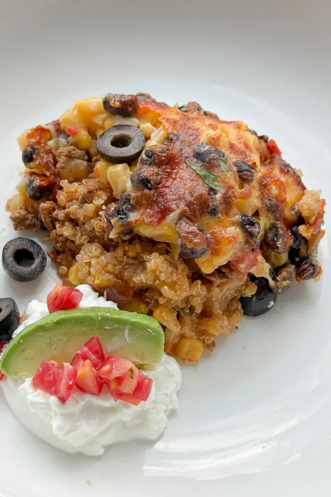 A slice of Mexican quinoa casserole on a plate next to Greek yogurt, avocado and diced tomatoes.