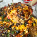 A close-up of a big spoonful of the Mexican quinoa casserole (on a wooden spoon) being held over the casserole on a counter.