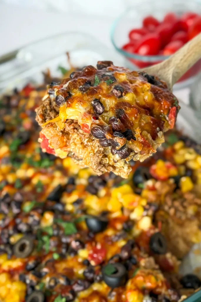 A close-up of a big spoonful of the Mexican quinoa casserole (on a wooden spoon) being held over the casserole on a counter.