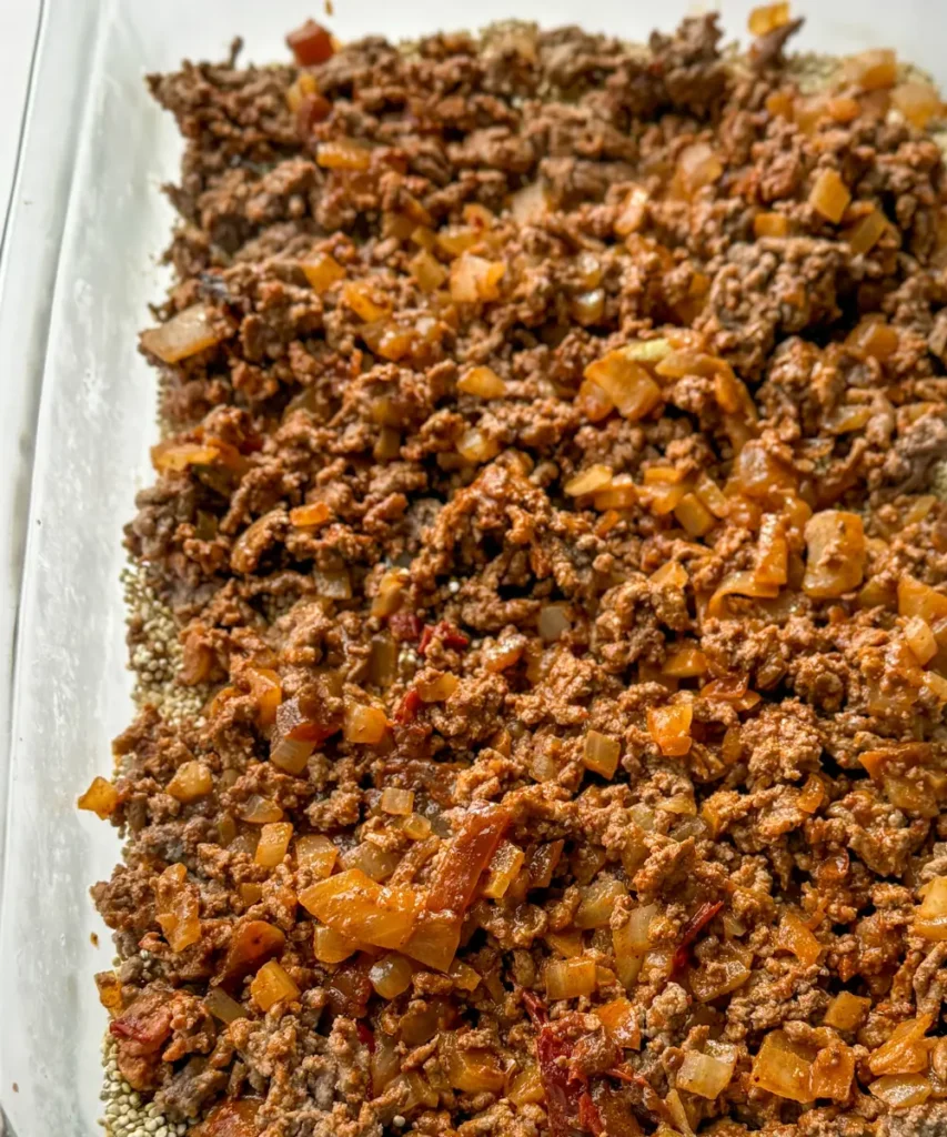 A casserole dish with raw quinoa and the ground beef layered in it.