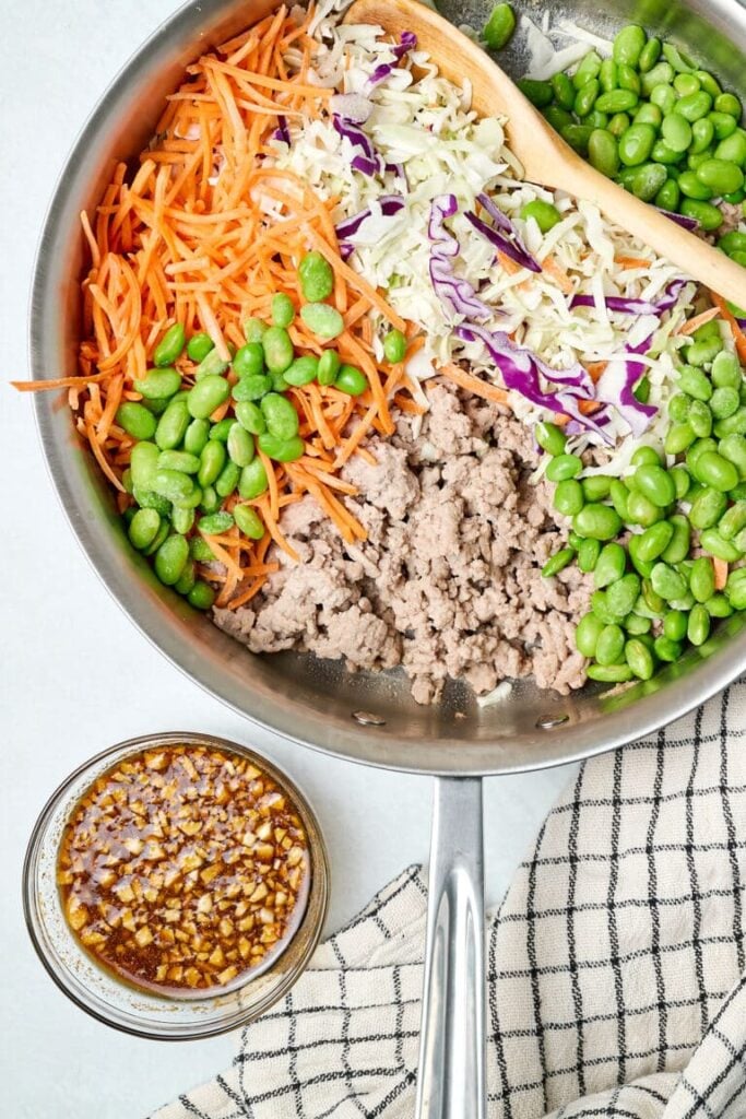 A birds-eye view of a pan of ground turkey, shelled edamame, sliced carrots, and veggies in a pan next to a bowl of sauce.