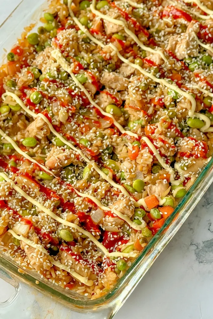 A birds-eye view of a fully assembled chicken fried rice casserole topped with spicy mayo and sesame seeds on a counter. 