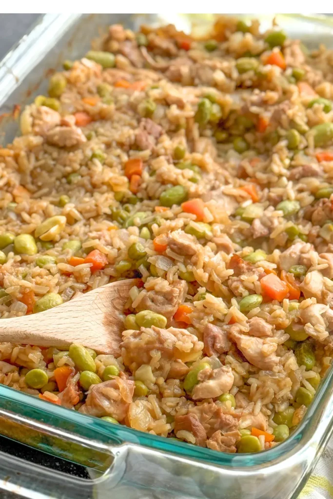 A baked chicken fried rice casserole in a glass casserole dish with a wooden spoon in the middle.