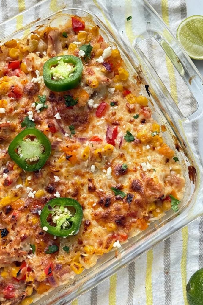 A birds-eye view of a baked street corn casserole on a decorative towel on the counter next to limes.