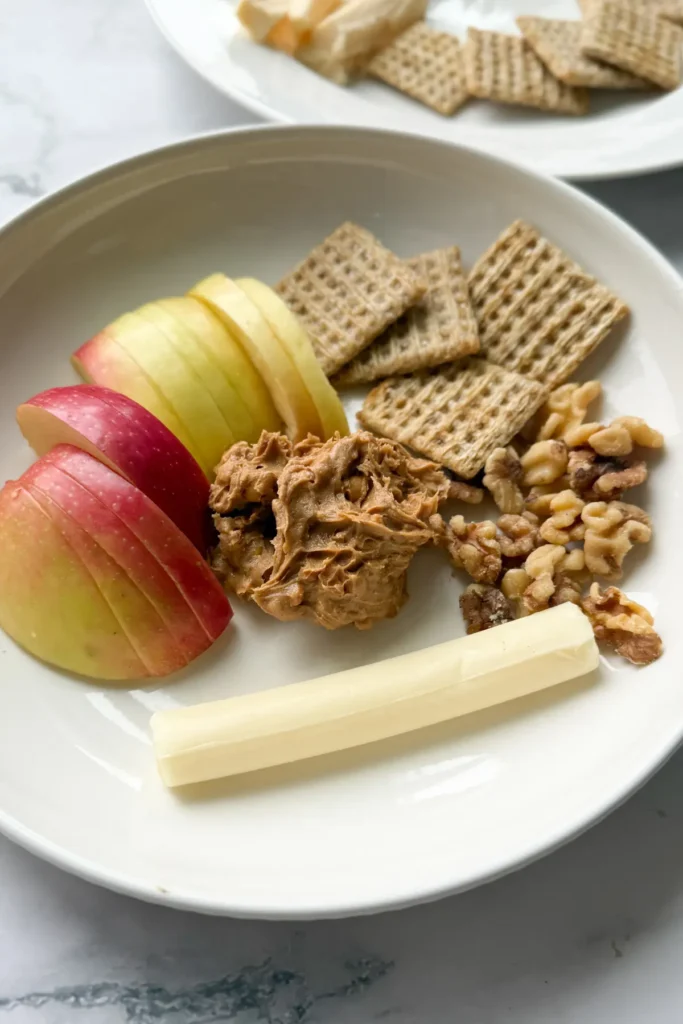 A snack plate with apple slices, a mozzarella cheese stick, chopped walnuts and Triscuit crackers on it.