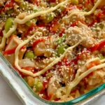 A close-up of a fully assembled chicken fried rice casserole topped with spicy mayo and sesame seeds on a counter.
