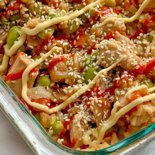 A close-up of a fully assembled chicken fried rice casserole topped with spicy mayo and sesame seeds on a counter.