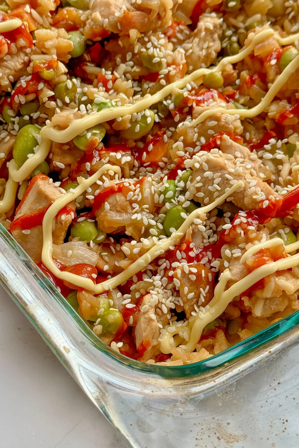 A close-up of a fully assembled chicken fried rice casserole topped with spicy mayo and sesame seeds on a counter.