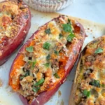 Three sweet potatoes stuffed with ground beef, cherry tomatoes, and onions topped with cheese after baking on parchment paper on a counter.