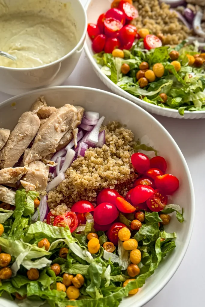 Two bright and beautiful fully assembled Caesar bowls without the dressingt on them next to a big bowl of healthy yogurt Caesar drizzle.