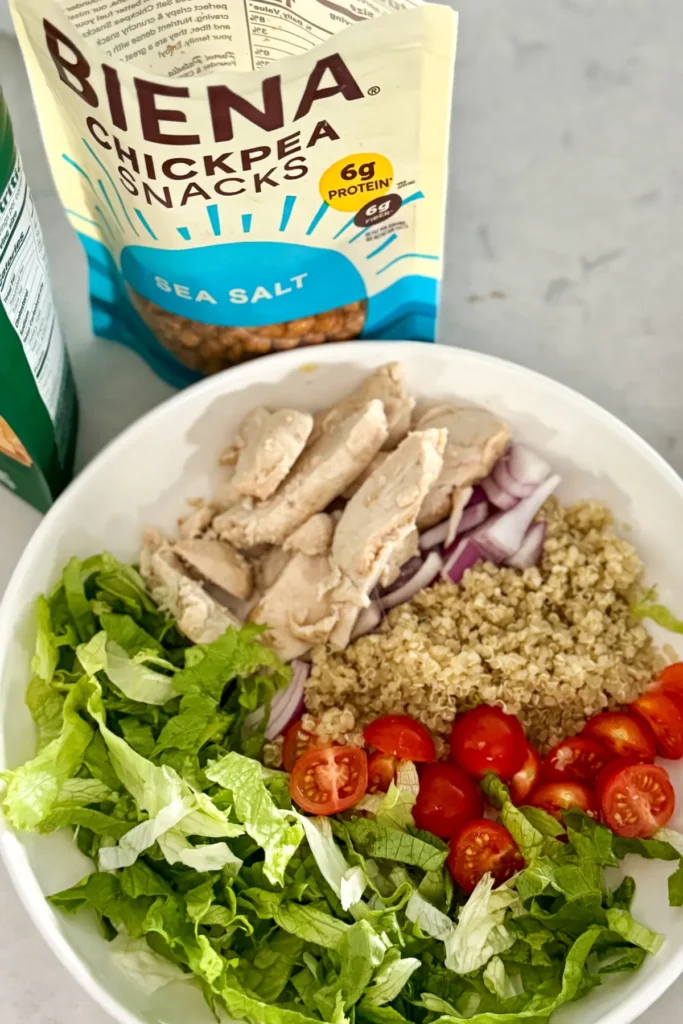 A bowl with shredded lettuce, tomatoes, chicken and quinoa in it next to a bag of store-bought roasted chickpeas on a counter.