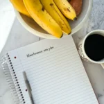 A pad of paper with "maintenance plan" writter on top next to a cup of coffee and a fruit bowl with bananas on a counter.