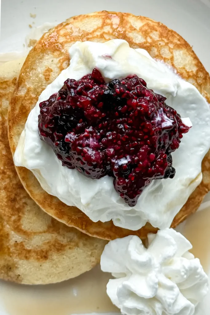 A close-up of pancakes stacked on top of one another on a white plate topped with yogurt and chia seed jam.