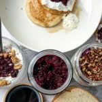 A birds-eye view of a jar of chia seed jam, PB and jelly sandwich, a stack of pancakes topped with yogurt, a yogurt parfait, and a cottage cheese bowl topped with walnuts on a counter.