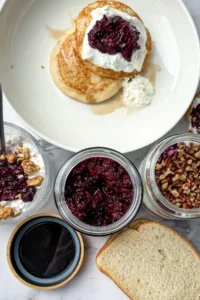 A birds-eye view of a jar of chia seed jam, PB and jelly sandwich, a stack of pancakes topped with yogurt, a yogurt parfait, and a cottage cheese bowl topped with walnuts on a counter.