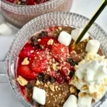 A birds-eye view of a fully assembled blended chia pudding topped with strawberries, marshmallow cream, mini marshmallows and chocolate chips in a glass bowl with a gold spoon in it.