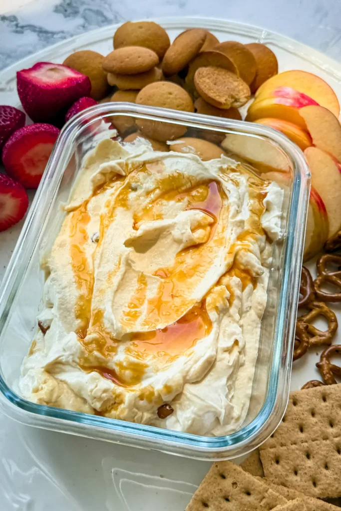 A close-up of easy salted caramel cheesecake dip in a meal prep storage container topped with caramel sauce surrounded by apples, vanilla wafers, and graham crackers.