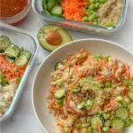 An avocado chicken and rice bowl next to assembled meal prep containers, half an avocado, and the sauce in a glass bowl.