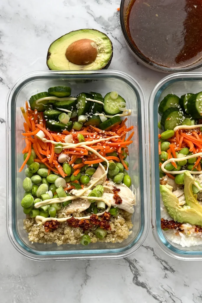 Two chicken, edamame, and rice meal prep containers on a counter next to a bowl of the Asian-inspired sauce and an avocado.