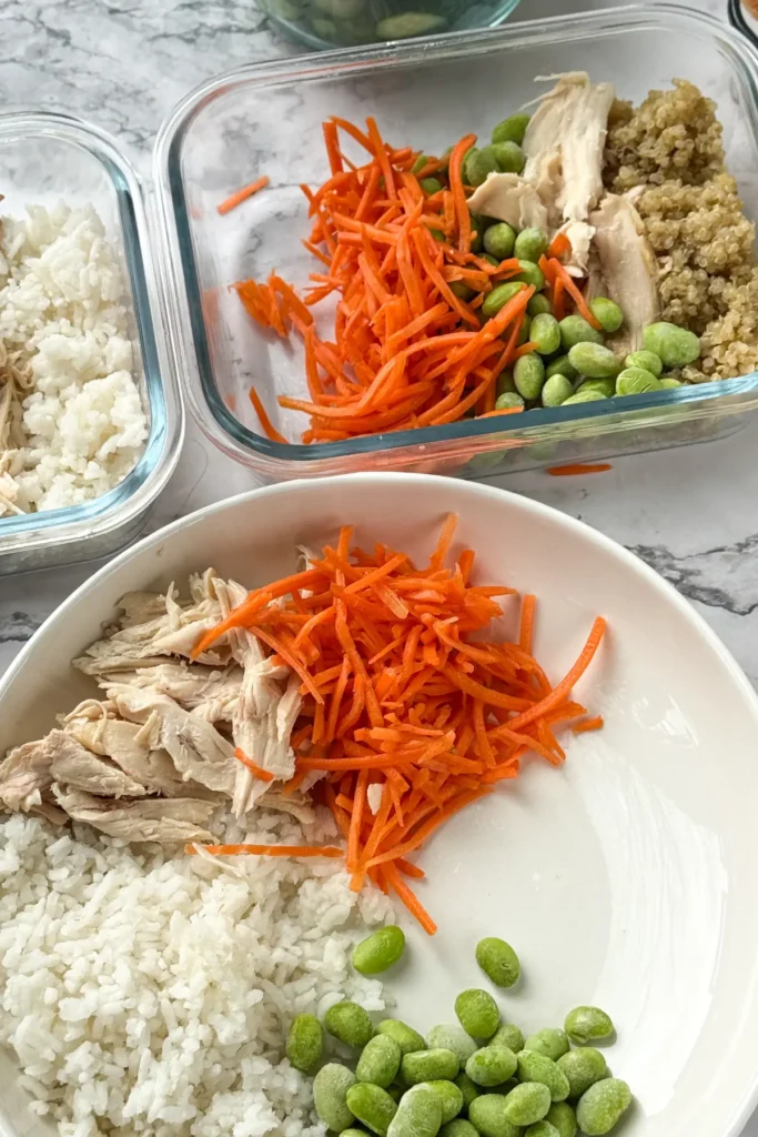 Assembling a bowl: rice, edamame, carrots and chicken on a white plate.