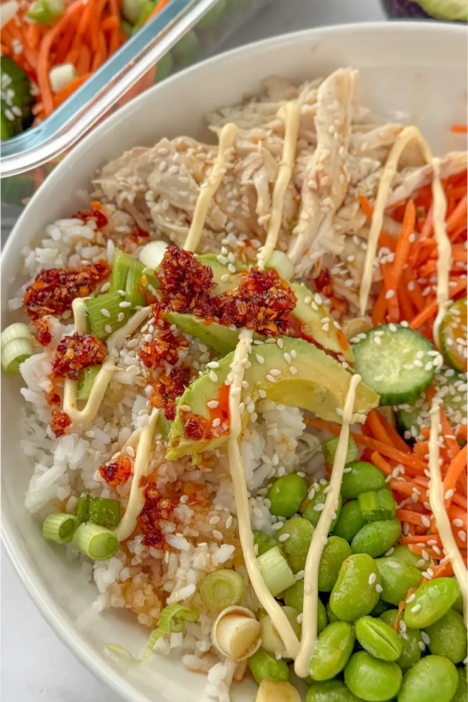 A close-up of a chicken and rice bowl topped with avocado, sesame seeds, chili crunch, and kewpie mayo.