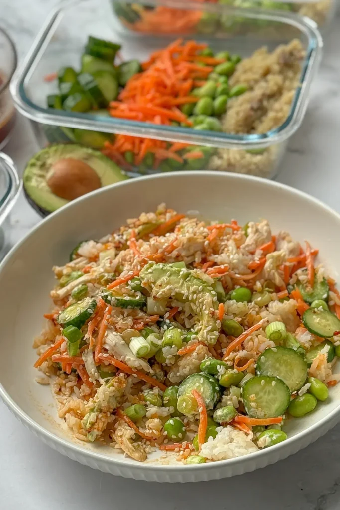 A chicken, rice, avocado, cucumber bowl topped with avocado and sesame seeds next to meal prep containers on a table.
