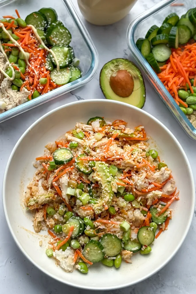 A birds-eye view of a chicken and rice bowl and two meal prep containers on a counter.