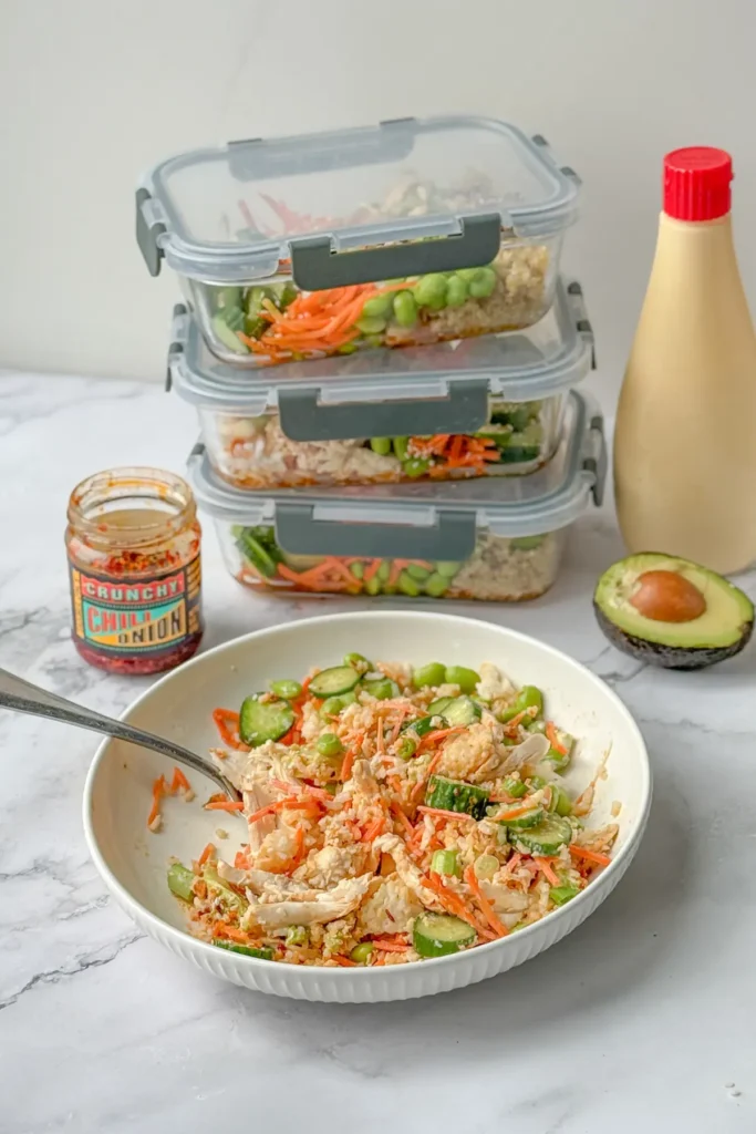 A half-eaten bowl next to a stack of meal prep containers on a counter next to chili crunch and kewpie mayo.