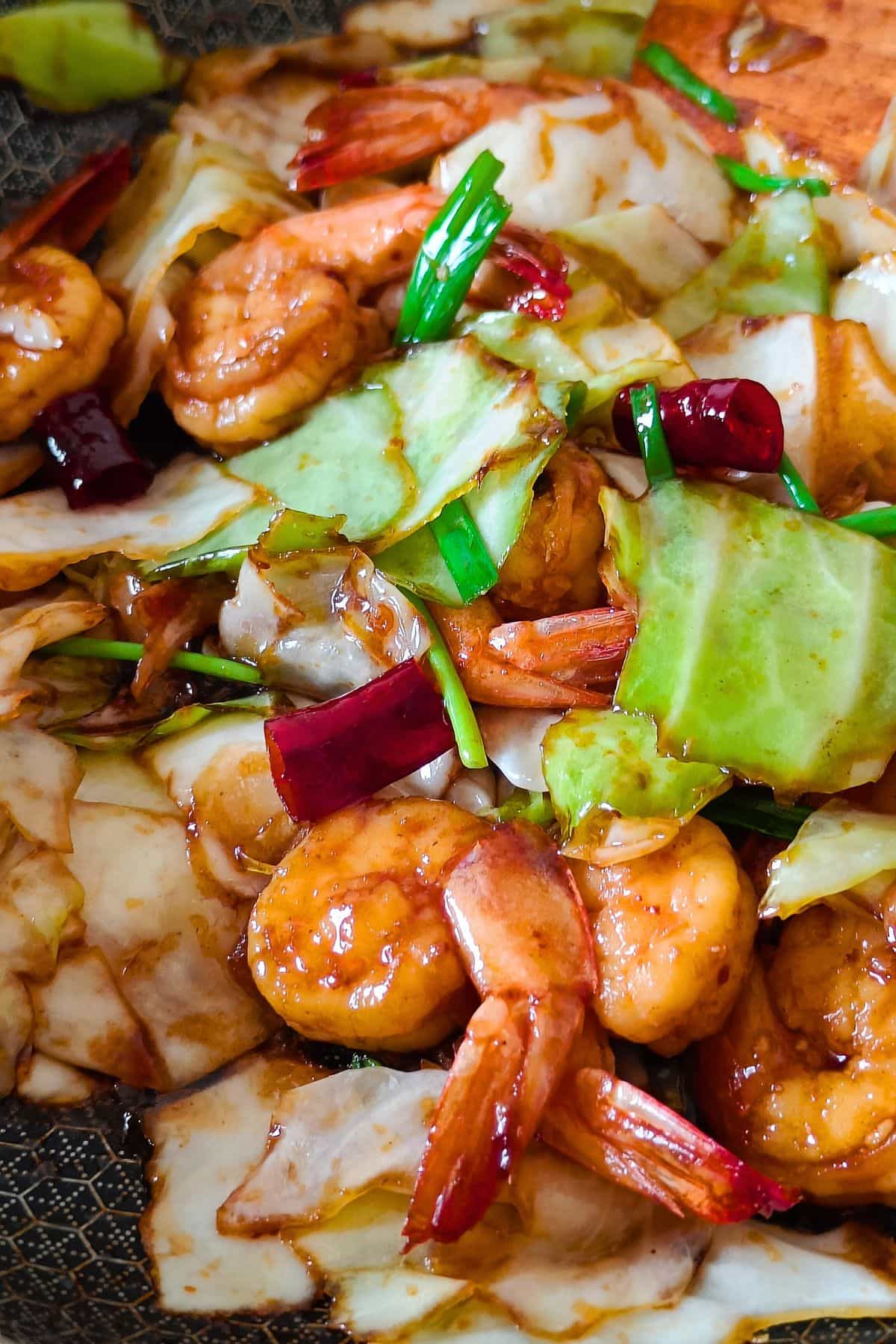 A close-up of a big pot of shrimp and cabbage stir-fry being stirred by a wooden spoon.